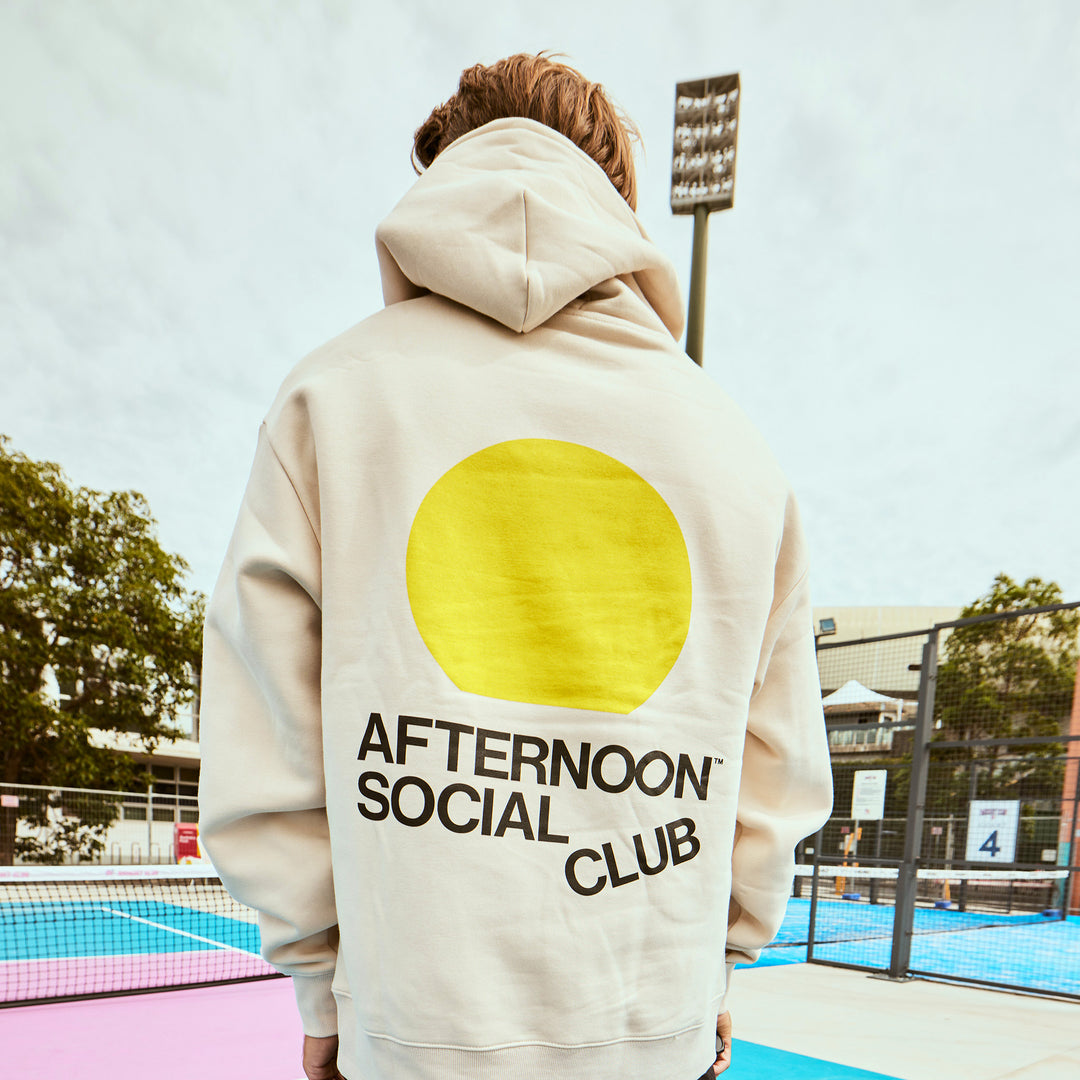 Man wearing ecru heavy hoodie with yellow sunset circle and Afternoon Social Club lettering on back.