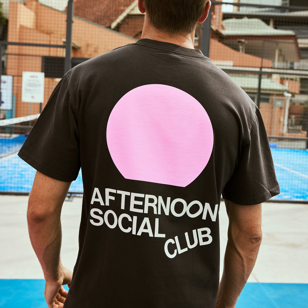 Back view of a man wearing a black Afternoon Social Club heavy tee featuring a pink graphic design.