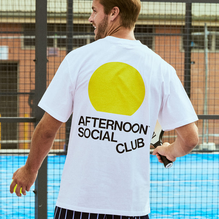 white Heavy Tee with yellow sunset circle and Afternoon Social Club lettering on back playing Pickleball.