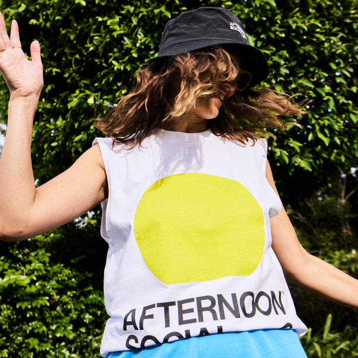Female having fun wearing white tank top with yellow sunset circle and Afternoon Social Club lettering on front.