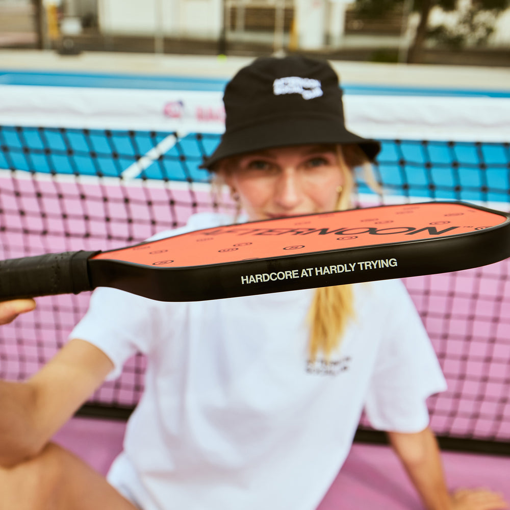 Female holding organge fiberglass pickleball paddle featuring the words hardcore at hardly trying on the side