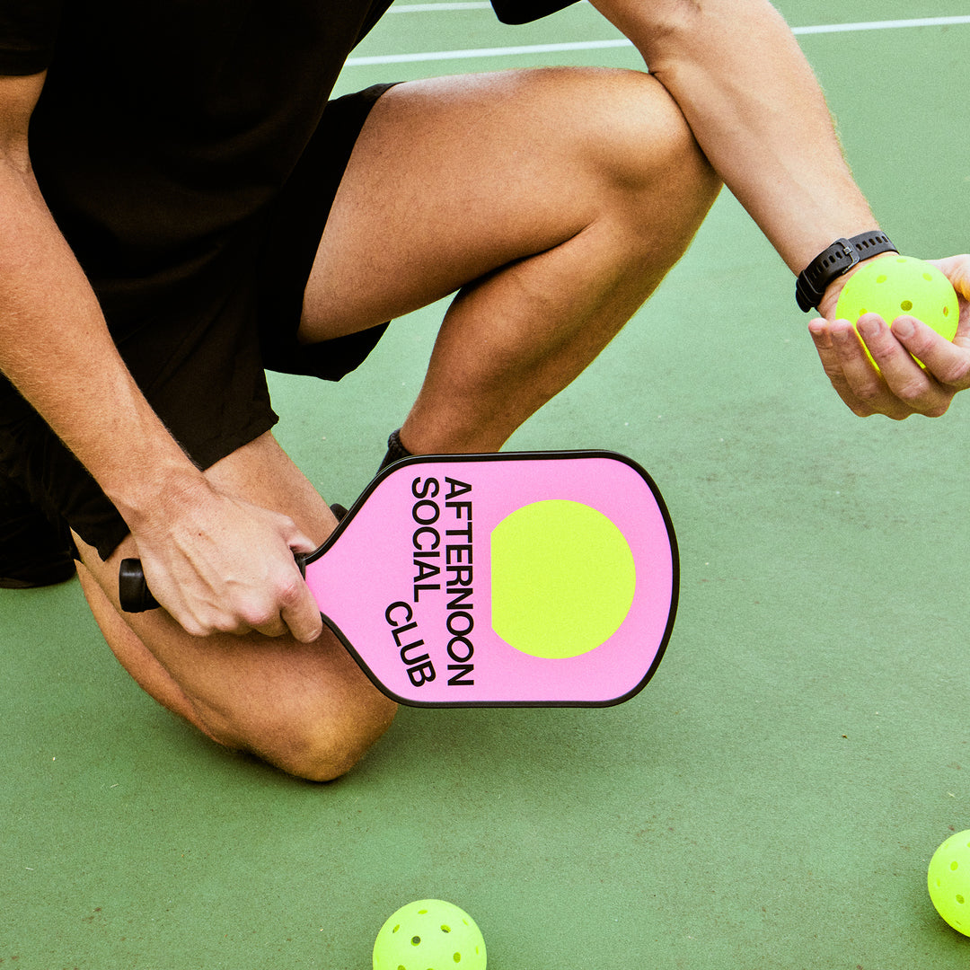 Model on the pickleball court holding Pink Fiberglass Pickleball Paddle featuring Afternoon Social Club logo and yellow sun